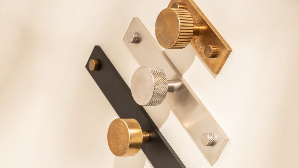 Three metallic door handles arranged diagonally on a white wall. The top is a gold handle with a textured, circular knob. The middle is a silver handle with a smooth, circular knob. The bottom is a black handle with a textured, circular knob.