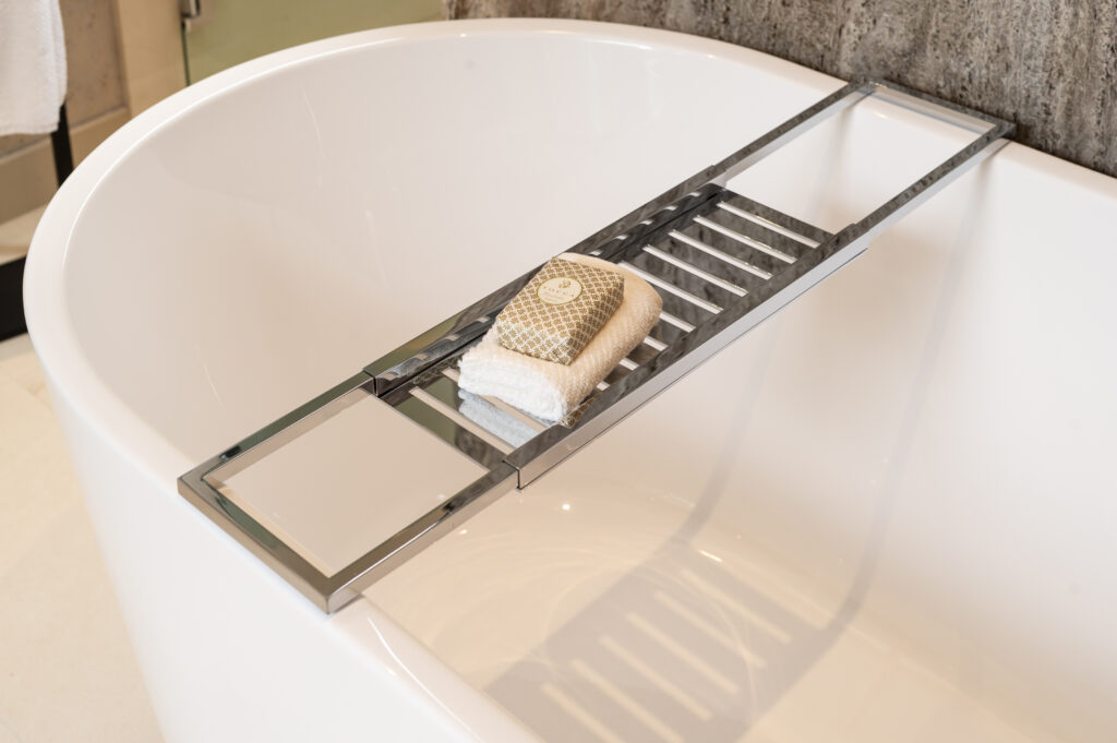 A modern white bathtub with a metal bath tray across it, holding a folded white towel, a woven washcloth, and a bar of soap. The tray has slats and a reflective surface on one end. The background includes a stone wall.