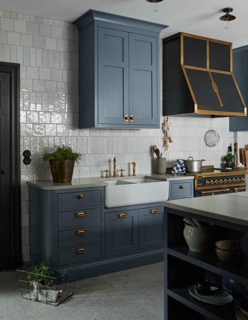 A kitchen with dark blue cabinets, a farmhouse sink, and brass fixtures. A large brass-trimmed range hood is above a stove. The backsplash features white tiles. Potted plants and kitchenware are neatly arranged. The countertop is a light stone color.