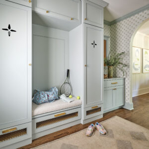 A cozy mudroom with a built-in bench, tennis racket, and balls. Light blue cabinets with decorative cutouts surround the space. A patterned rug and sneakers are on the wooden floor. A doorway leads to a wallpapered hallway with framed art.