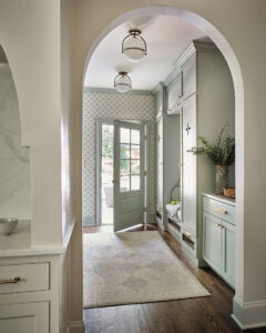 A hallway with pale green cabinets and built-in storage leads to a door opening to a garden. The space has wooden flooring, a rug, and patterned wallpaper. Two ceiling lights hang above, and a plant decorates the counter.