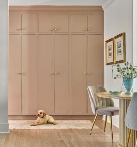 A small poodle lies on a light rug in front of a wall of pinkish cabinets with gold handles. To the right, there's a round table with a beige chair and a vase of flowers. Two framed pictures hang above the table on a white wall.
