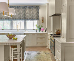 A bright kitchen with light cabinetry and a marble island. A window with a patterned blind lets in natural light. A stove is topped with a pot, and a vase with pink flowers sits on the counter. A fruit bowl and stool are near the island.