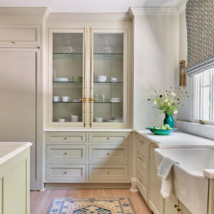 A light, elegant kitchen features a glass-door cabinet with dishes, a farmhouse sink with a decorative towel, flowers in a green vase, and a nearby window with patterned curtains. The wooden floor has a decorative rug.