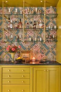 A home bar with mustard yellow cabinets and drawers, topped with a candle and vase of flowers. Glass shelves display various bottles and barware. The background features a colorful floral and geometric patterned wallpaper.