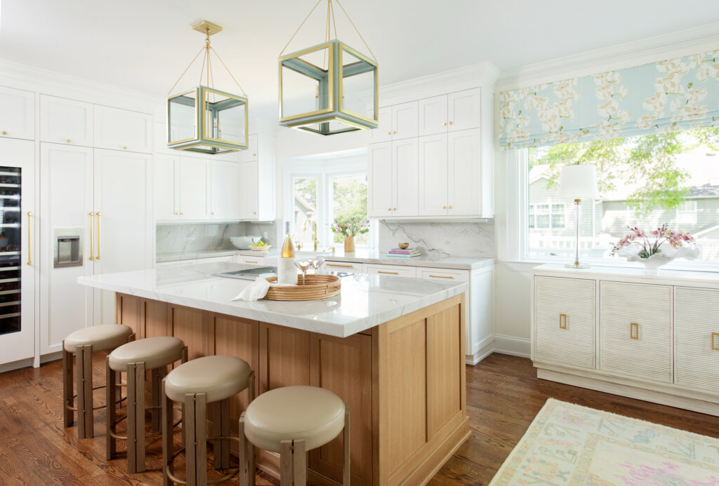 Spacious kitchen with white cabinets, wood accents, and a large central island featuring light wood and a white countertop. Four round stools surround the island. Two modern pendant lights hang above, and a window with floral curtains allows natural light in.