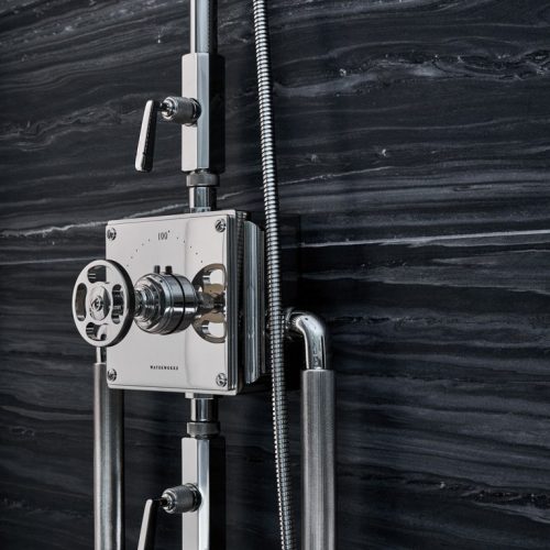 Close-up of a modern shower panel with a metallic finish against a dark, textured stone wall. The panel features a central dial and multiple levers for water adjustment, alongside sleek, vertical pipes and a flexible hose.