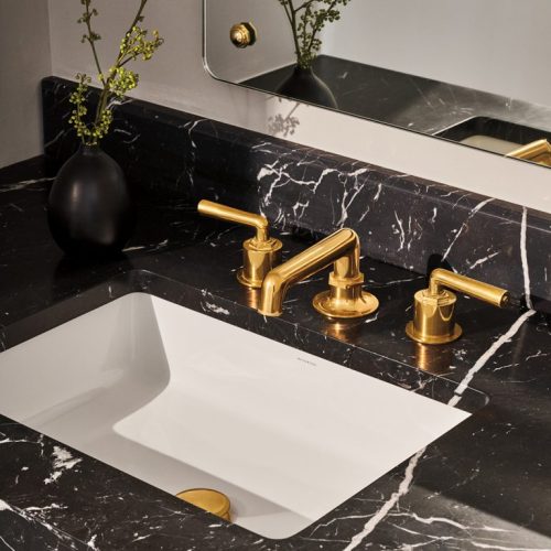 A modern bathroom sink with a black marble countertop features a gold faucet. A small black vase with green sprigs rests beside the sink. A rectangular mirror is mounted above.