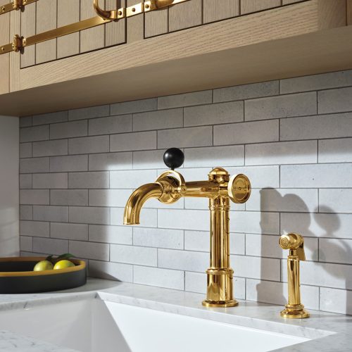 A modern kitchen sink with a gold faucet and matching hardware, set against a light gray tiled backsplash. Marble countertop holds a bowl of assorted fruits. Wooden cabinetry with gold accents is seen above.