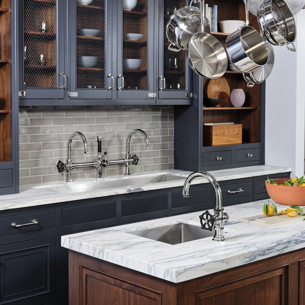 A modern kitchen with dark cabinetry and a marble countertop. There's a silver faucet on a central island, and another faucet along the wall with a backsplash. Pots hang from a ceiling rack, and open shelves display dishes.