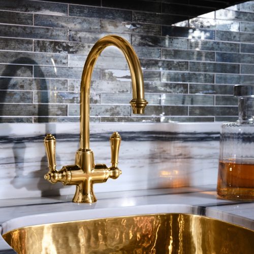 Luxurious kitchen faucet in polished gold with an ornate design, accompanied by a gold sink. The backdrop features reflective, dark-toned tiles, and a glass container with amber liquid rests nearby on the marbled countertop.