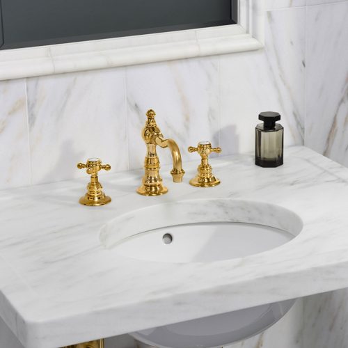 A luxurious bathroom sink with a marble countertop features elegant gold faucets and a matching gold drain pipe. A small, dark glass bottle is placed beside the faucet against a marble wall background.