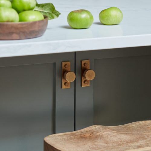 A kitchen scene featuring a wooden stool, green cabinet doors with round brass knobs, and a white countertop. A wooden bowl with green apples and two loose apples sit on the countertop.