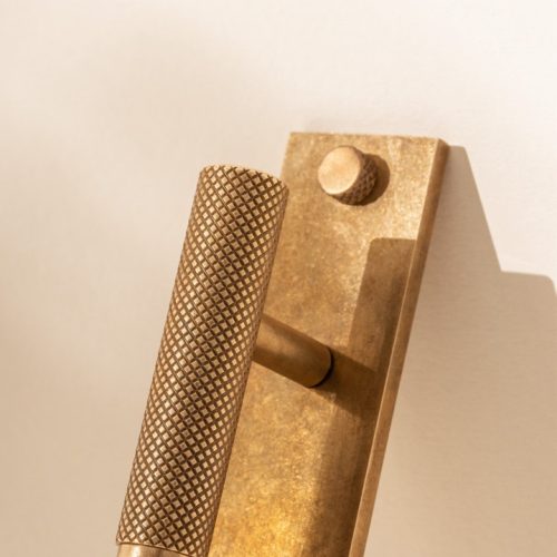 Close-up of a modern, textured brass door handle mounted on a light-colored door. The handle has a cylindrical shape with a knurled grip, casting a shadow to the right.
