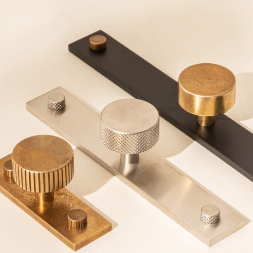 Three cylindrical door knobs on rectangular bases are displayed against a neutral background. The knobs vary in texture and color: one is gold with ridges, another is silver with a smooth finish, and the last is matte black.