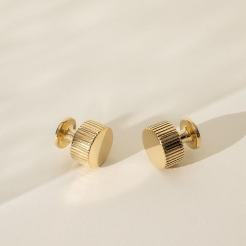 A pair of gold stud earrings with a ribbed texture rests on a light-colored surface. Soft shadows are cast, suggesting gentle lighting from one side.