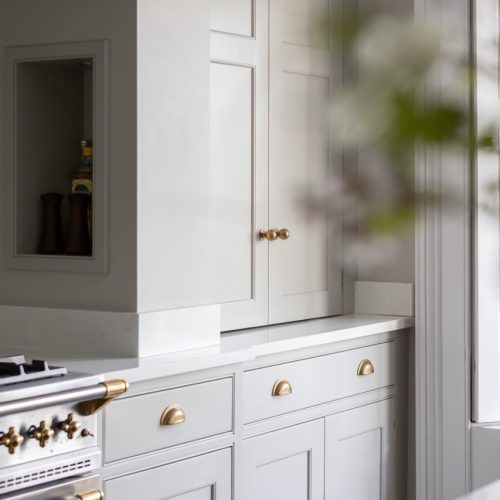 White kitchen cabinets with brass handles and knobs, next to a white oven with brass accents. A small alcove holds jars, and blurred greenery is visible on the right side of the image.