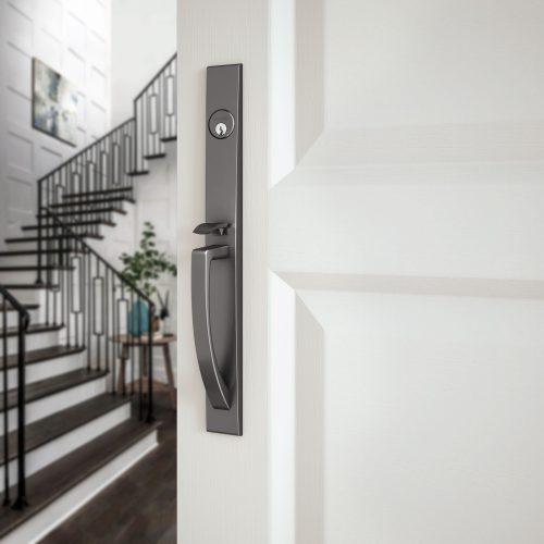 Close-up of a modern, dark door handle and lock on a white door. The door is ajar, revealing a staircase and a framed picture in a well-lit hallway.