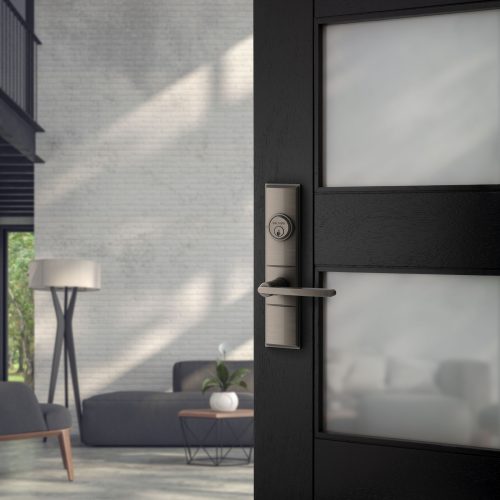 A modern interior features a black door with a sleek metal handle partially open, revealing a room with white brick walls, a gray sofa, a white floor lamp, and a round coffee table with a white orchid. Sunlight streams in, casting soft shadows.