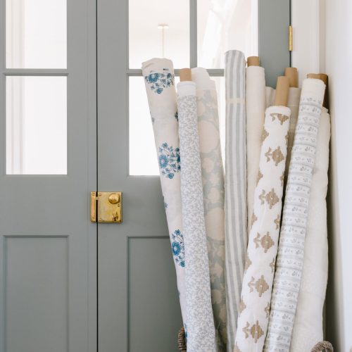 A woven basket filled with various rolled wallpaper stands beside light gray paneled double doors with glass panels. The wallpapers feature different patterns, including floral, geometric, and stripes in shades of blue, beige, and white.