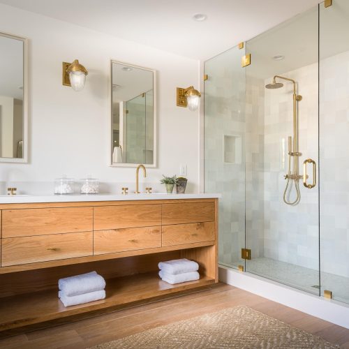 Modern bathroom with a wooden double vanity, gold fixtures, and two mirrors. A glass-enclosed shower with a wooden stool and gold showerhead is on the right. The space is decorated with potted plants and rolled towels on an open shelf.