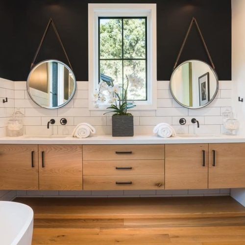 A modern bathroom with a double vanity featuring wooden cabinets. Two round mirrors hang above the sinks, and a potted orchid sits in the center. The walls are white-tiled with a black upper section. Natural light enters through a window.