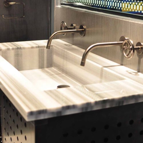 A modern bathroom with a rectangular marble sink featuring two wall-mounted faucets. The wall is decorated with a patterned glass panel above the sink. The cabinet below has a perforated metal design.