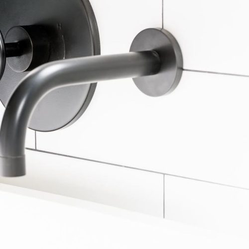 Close-up of a modern black matte wall-mounted faucet and handle against a white tiled wall. The design is minimalist with a circular backplate and a curved spout extending outward.