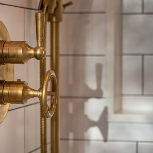 Close-up of vintage brass shower controls on a white tiled wall. The design features a round backplate with two metal levers and an ornate circular handle casting shadows on the tiles.