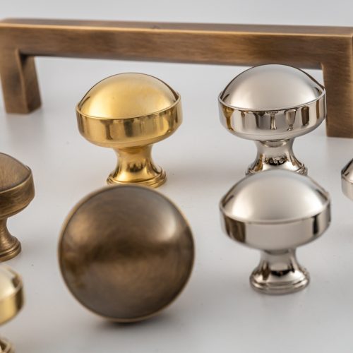 A collection of metallic cabinet knobs and handles on a white surface. The knobs are in various finishes, including brass, bronze, and silver, with different shapes such as round and square, alongside rectangular handles.