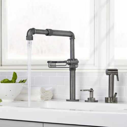 A modern kitchen faucet with water flowing into a white sink. A small bowl of basil and folded towels are on the counter. The backsplash is white subway tile, and a window is in the background.