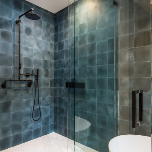 A modern bathroom features a glass-enclosed shower with dark blue textured tiles, a ceiling-mounted rain showerhead, and a handheld shower. Natural light enters through a small skylight above, illuminating the white tile floor.