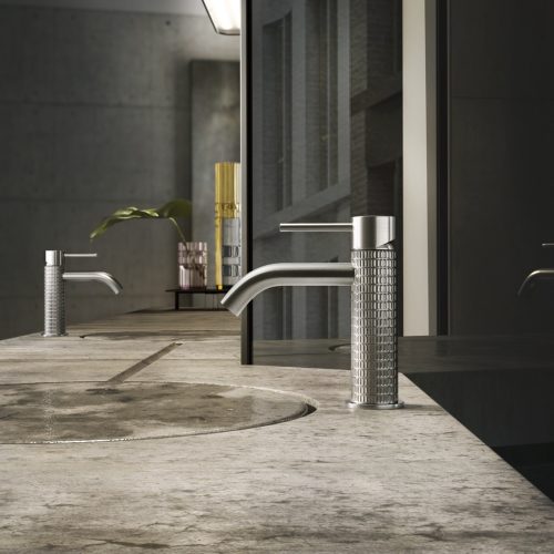 A modern bathroom sink with a concrete countertop and a sleek, cylindrical metal faucet. A minimalistic mirror reflects the room. In the background, a decorative plant and a gold-toned object sit on the counter. The wall is grey and textured.