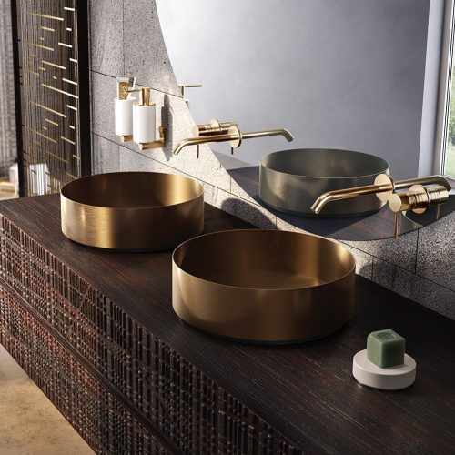 A modern bathroom with two circular, gold-colored sinks on a dark textured wooden countertop. Minimalist gold faucets extend from the wall. A large mirror with rounded corners reflects the room. A small green soap on a dish is on the counter.