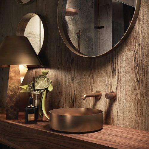 A modern bathroom features a wooden countertop with a round, gold sink and wall-mounted faucet. A gold lamp with a textured base and a plant in a vase sit nearby. Two round mirrors are mounted on the wood-paneled wall.