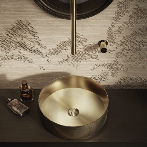 Luxurious bathroom sink with a round, gold basin on a dark countertop. A gold faucet extends from the ceiling. Toiletries in elegant containers are placed nearby. The wall has a textured, patterned finish. Mood lighting enhances the sophisticated ambiance.