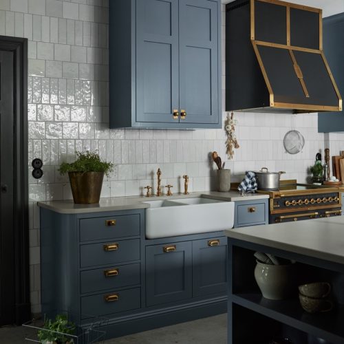 A stylish kitchen with blue-gray cabinets, a farmhouse sink, a brass-accented cooker hood, and a vintage stove. The countertop holds a potted plant and kitchenware. White tiled walls and brass handles complement the modern classic design.