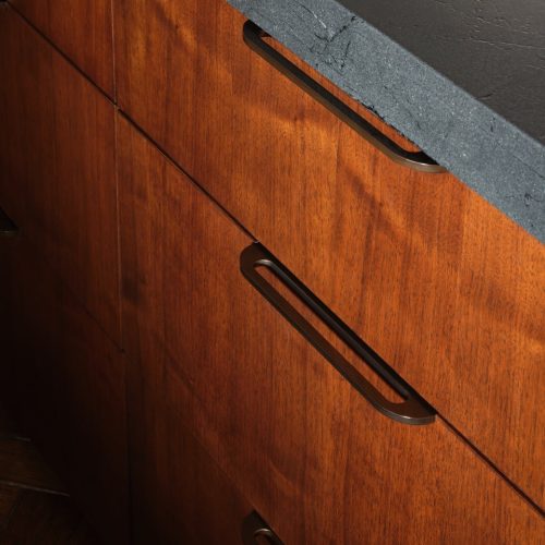 Wooden kitchen cabinets with sleek, dark metal handles below a textured black countertop. The warm wood grain contrasts with the countertop's matte finish, creating a modern and stylish appearance.
