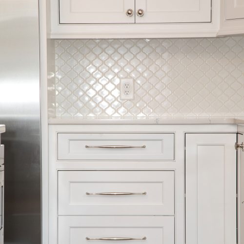 White kitchen cabinets with silver handles and a matching dishwasher. The backsplash features a glossy, overlapping fish scale tile design in white. There is a power outlet in the center of the backsplash.