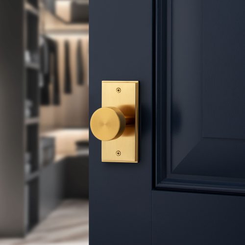 Close-up of a brass doorknob on a dark blue door, slightly ajar, revealing a blurred view of a modern walk-in closet with hanging clothes and shelves in the background.