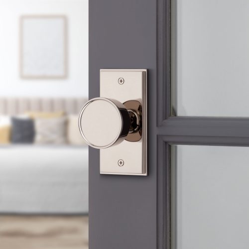 Close-up of a polished silver door knob on a gray door with a rectangular backplate. The door is slightly open, revealing a softly lit bedroom in the background with a bed, pillows, and a blurred window.