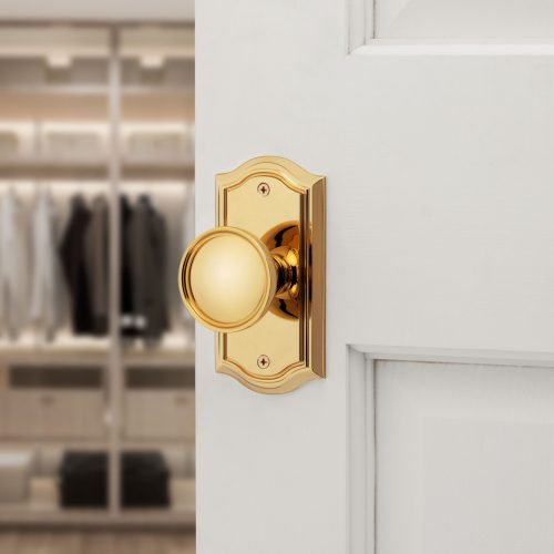 A polished brass doorknob with a decorative plate is mounted on a white door. The door is slightly ajar, revealing a blurred view of a closet with neatly hung clothes in the background.