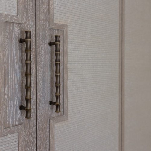 Close-up of two textured wooden cabinet doors with ornate bronze handles designed to resemble bamboo. The handles are vertically aligned on each door, which features a geometric panel design. A similar door is partially visible on the right.