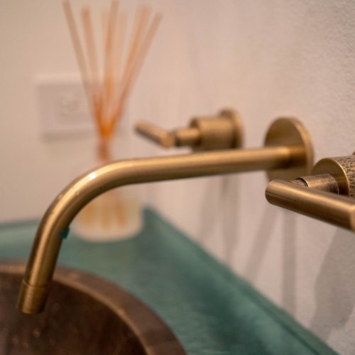 Close-up of a brass bathroom faucet and handles mounted on a white wall. The faucet extends over a green countertop with a copper-colored sink. In the blurred background, a set of reed diffusers is visible.