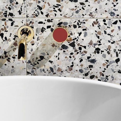 Close-up of a terrazzo bathroom wall with a gold faucet and a red circular control knob. A white curved bathtub edge is visible at the bottom of the image. The terrazzo pattern features gray, black, and brown speckles.