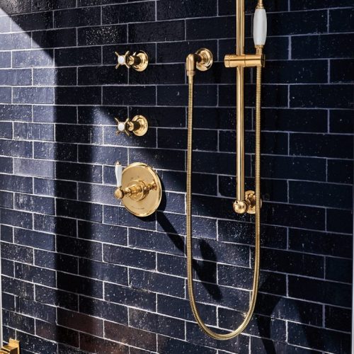 A luxurious shower setup with gold fixtures against a dark tiled wall. It features a handheld showerhead, controls, and a mounted folding wooden bench. Natural light highlights the shiny gold accents.