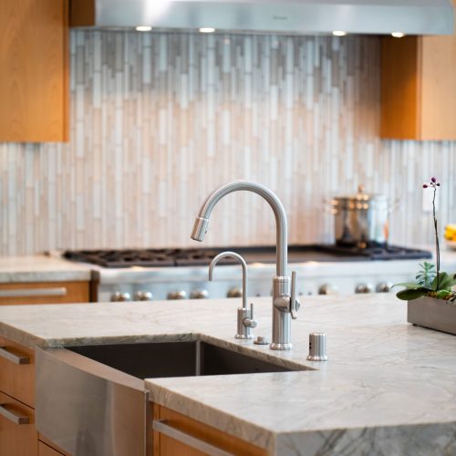 Modern kitchen with a marble countertop island featuring a stainless steel sink and curved faucet. Behind it, there's a stove with a metallic backsplash and wooden cabinets. A small potted plant sits on the counter.