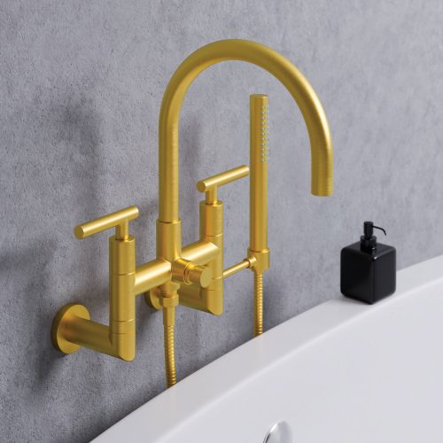 A modern gold bathroom faucet with a curved spout and dual handles is mounted on a gray wall above a white bathtub. A small, black soap dispenser is placed on the edge of the bathtub.