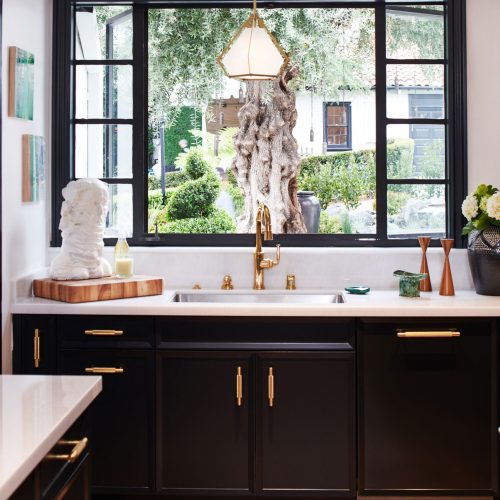 Stylish kitchen with black cabinets, gold hardware, and white countertops. Large window over the sink reveals a scenic garden with greenery. Geometric pendant light hangs above the sink. Parquet flooring adds warmth to the space.